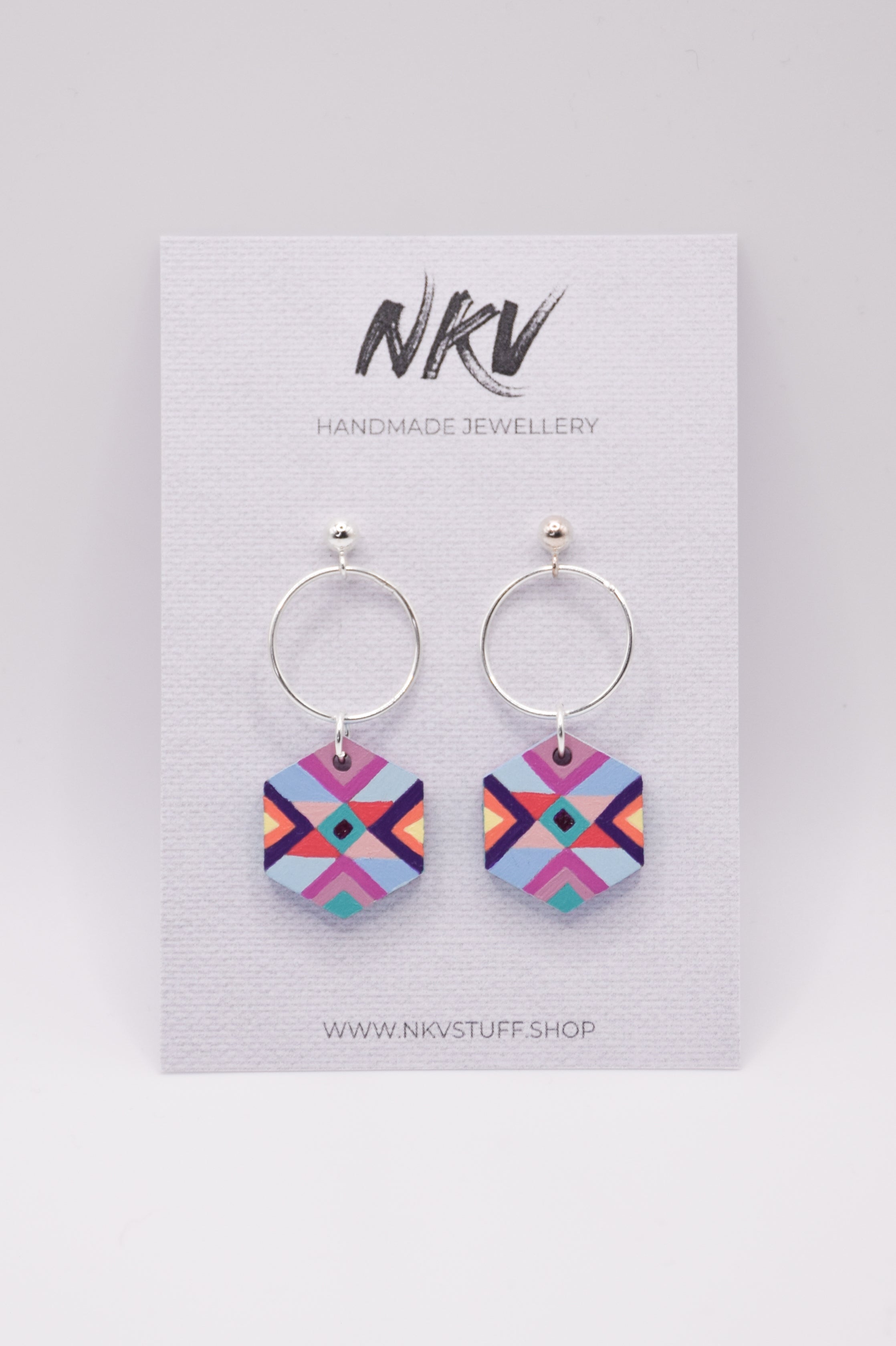Hexagonal hand-painted wooden earrings in purple and blue colours with silver hoops on a NKV branded card